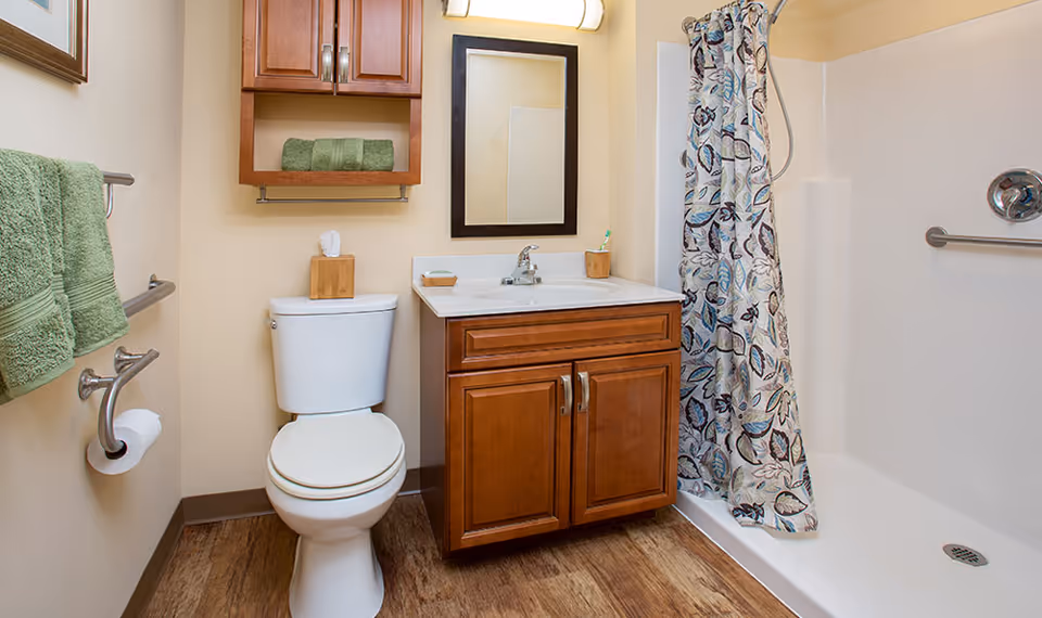 A clean and well-lit bathroom featuring a white toilet, a wooden vanity with a sink and mirror above it, a shower with a floral patterned curtain, green towels hanging on a towel rack, and a wooden cabinet mounted on the wall above the toilet.