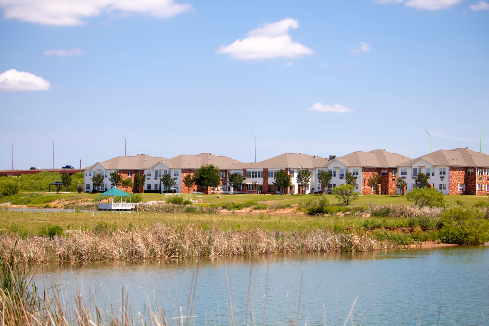 View of Elison Assisted Living of Lake Wellington building from across a pond with grassy area and a small gazebo near the water under a blue sky with a few clouds.