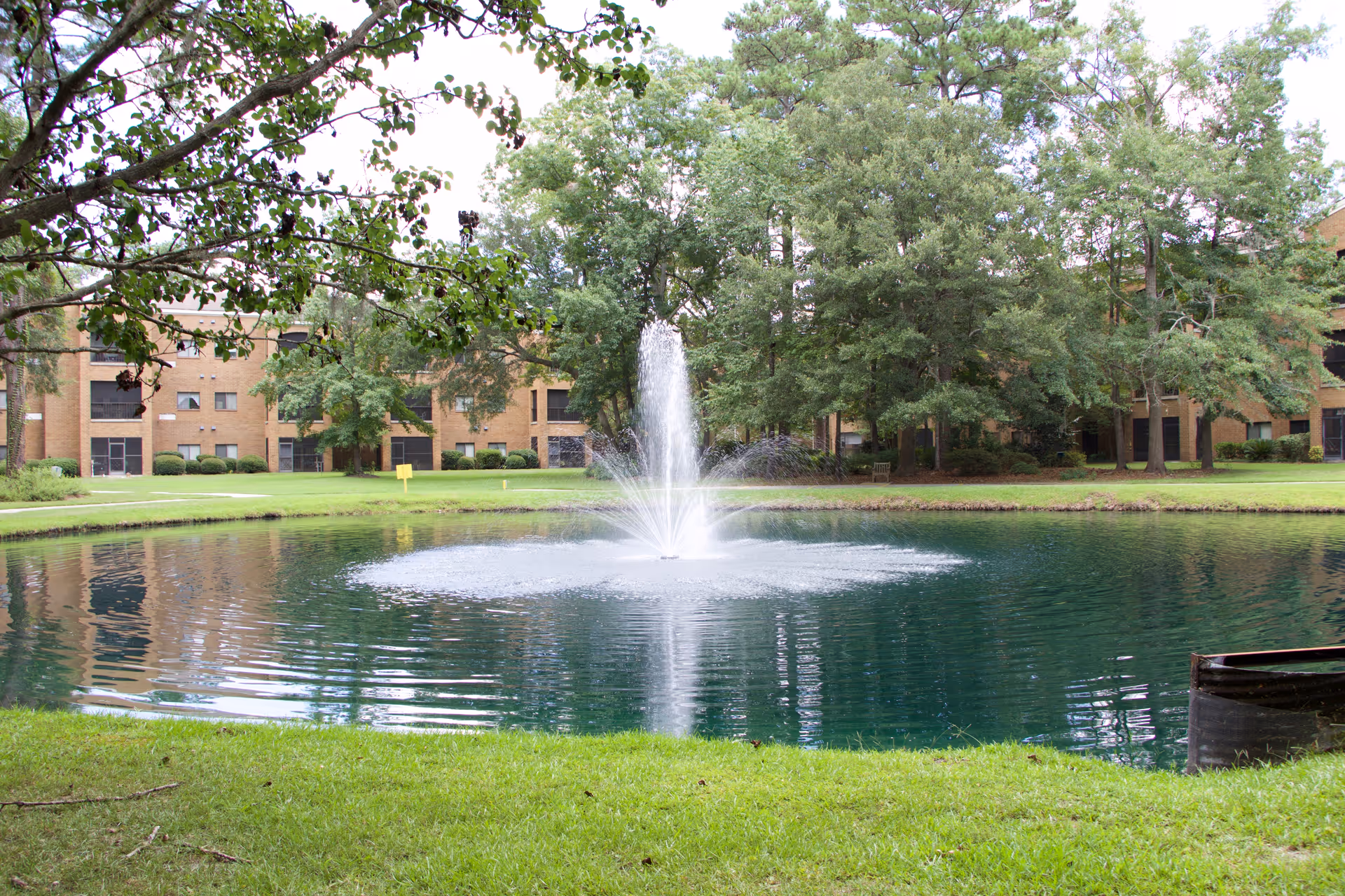 A serene outdoor scene featuring a pond with a central water fountain surrounded by green grass and trees. In the background, there is a multi-story brick building partially visible through the trees.