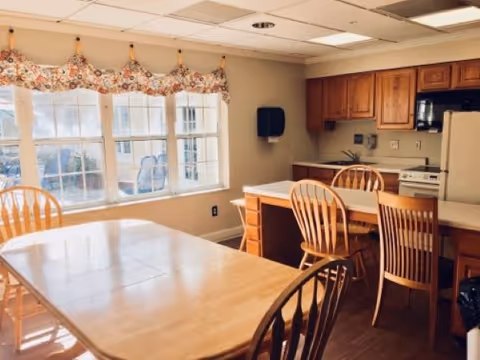 A bright kitchen and dining area with wooden chairs and tables. The kitchen features wooden cabinets, a white refrigerator, a microwave, and a stove. Large windows with floral valances allow natural light to fill the room.