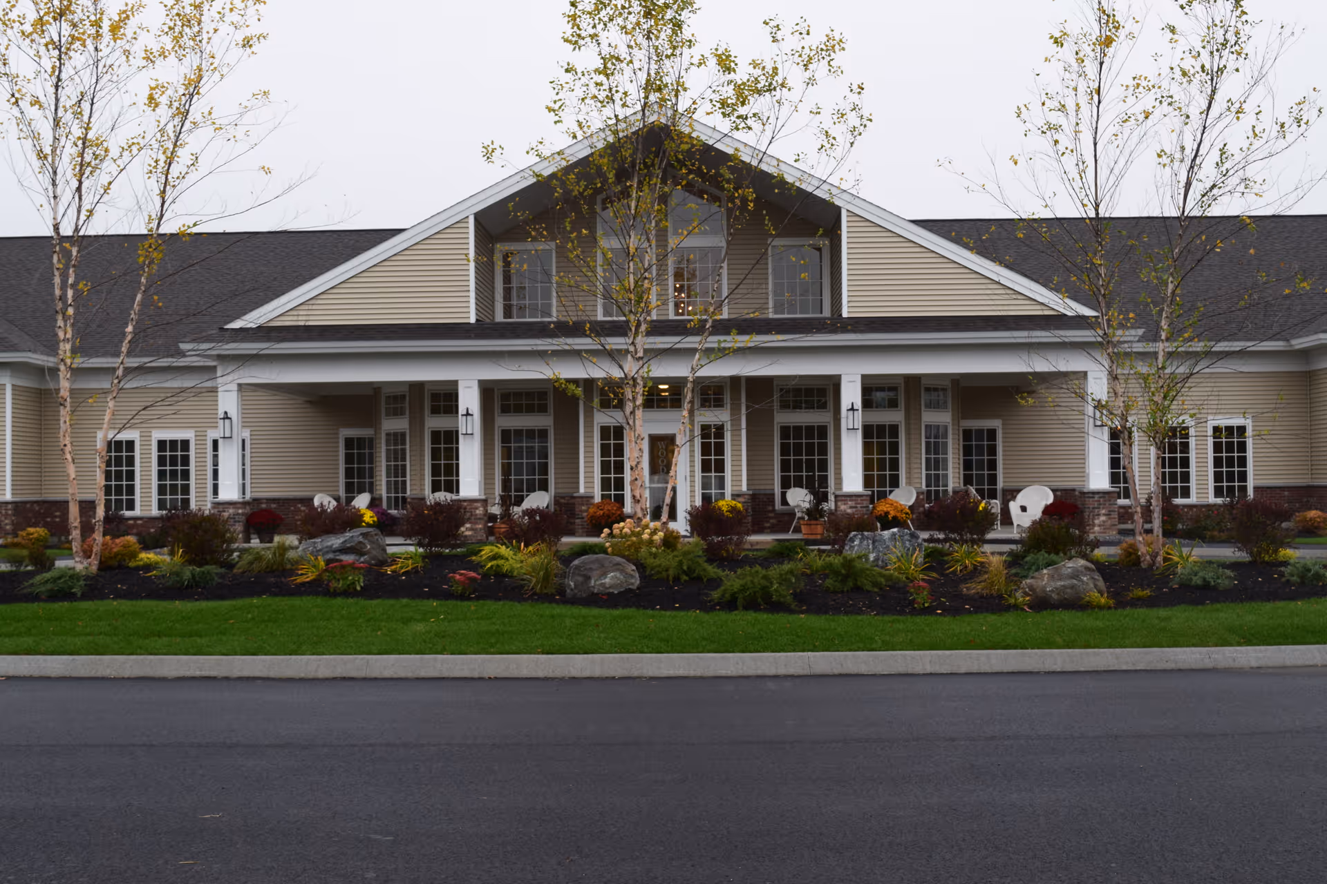 Front exterior view of a single-story building with beige siding and a dark roof, featuring a covered porch with white columns and several white chairs. The landscaped area in front includes small trees, shrubs, flowers, and large rocks, with a paved road in the foreground.