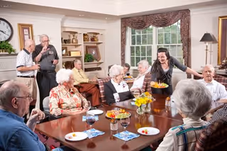 A group of elderly people sitting around a large dining table in a well-lit room with large windows and traditional decor. A caregiver is serving food to one of the residents. The table is set with plates of fruit and glasses of water or wine. The room has a cozy and welcoming atmosphere with framed pictures on the walls and a clock above a fireplace.
