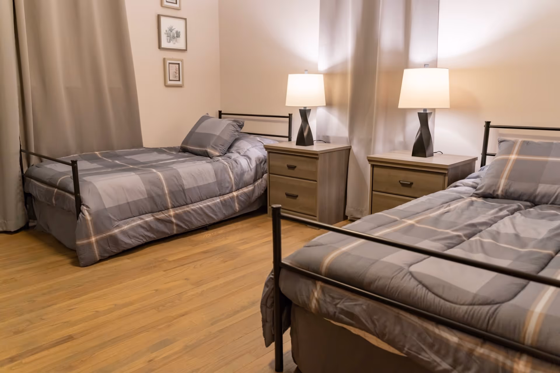 A bedroom with two single beds, each with gray plaid bedding. Between the beds is a wooden nightstand with two drawers, topped with a modern black lamp with a white lampshade. The room has light-colored walls, wooden flooring, and curtains covering the windows. Three small framed pictures hang on the wall above one of the beds.