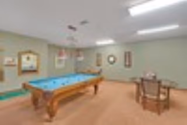 A recreational room with a pool table in the center, overhead lights hanging above it, framed pictures on the walls, and a small table with chairs in the corner.
