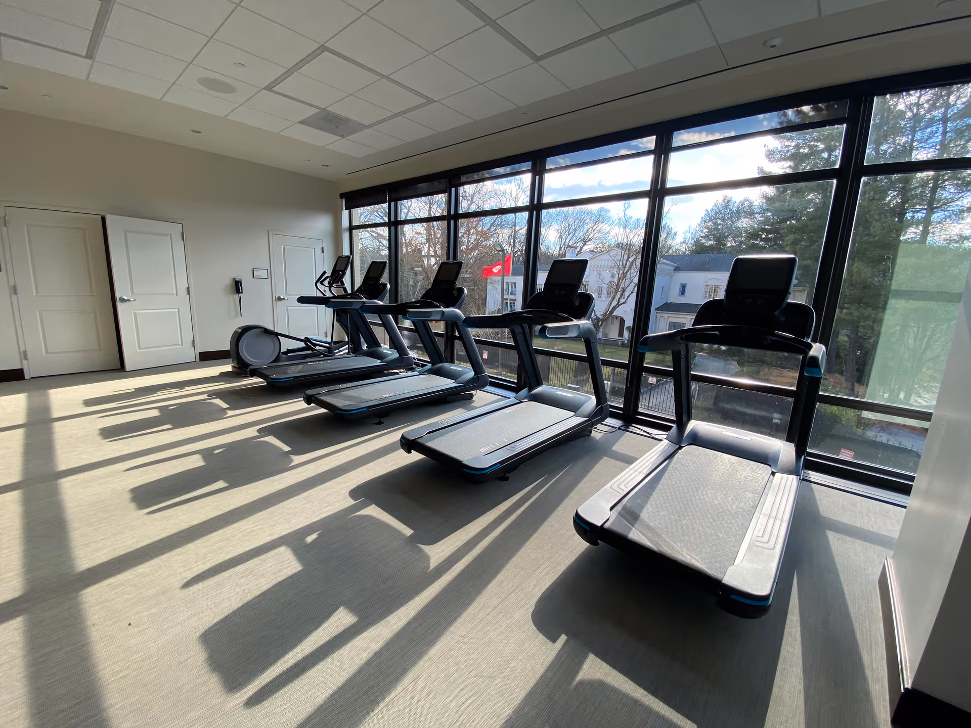 A bright fitness room with four treadmills lined up facing large floor-to-ceiling windows that offer a view of trees and buildings outside. The room has light-colored flooring and a white ceiling with recessed lighting.