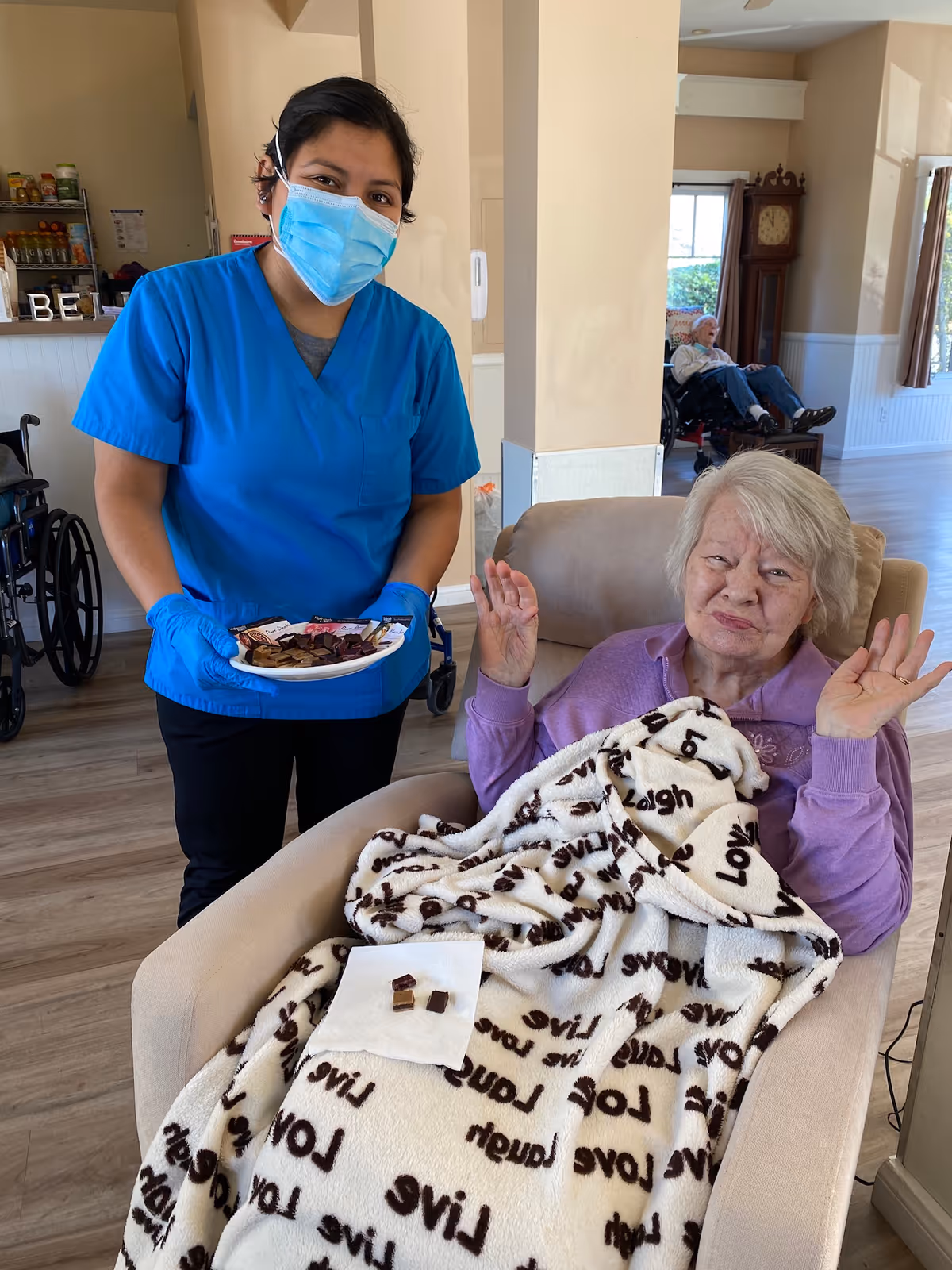 A healthcare worker wearing blue scrubs, a face mask, and gloves is standing next to an elderly woman sitting in a beige armchair covered with a white blanket that has words like 'Love' and 'Live' printed on it. The healthcare worker is holding a plate with small pieces of food, and the elderly woman is raising her hands with a slight smile. In the background, there is another elderly person in a wheelchair near a window and a tall grandfather clock.