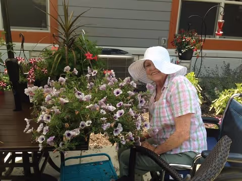 An elderly woman wearing a large white sunhat and a pink and white checkered shirt is sitting outdoors next to a table with a large pot of blooming purple and white flowers. The setting appears to be a garden or patio area with plants and a building wall in the background.