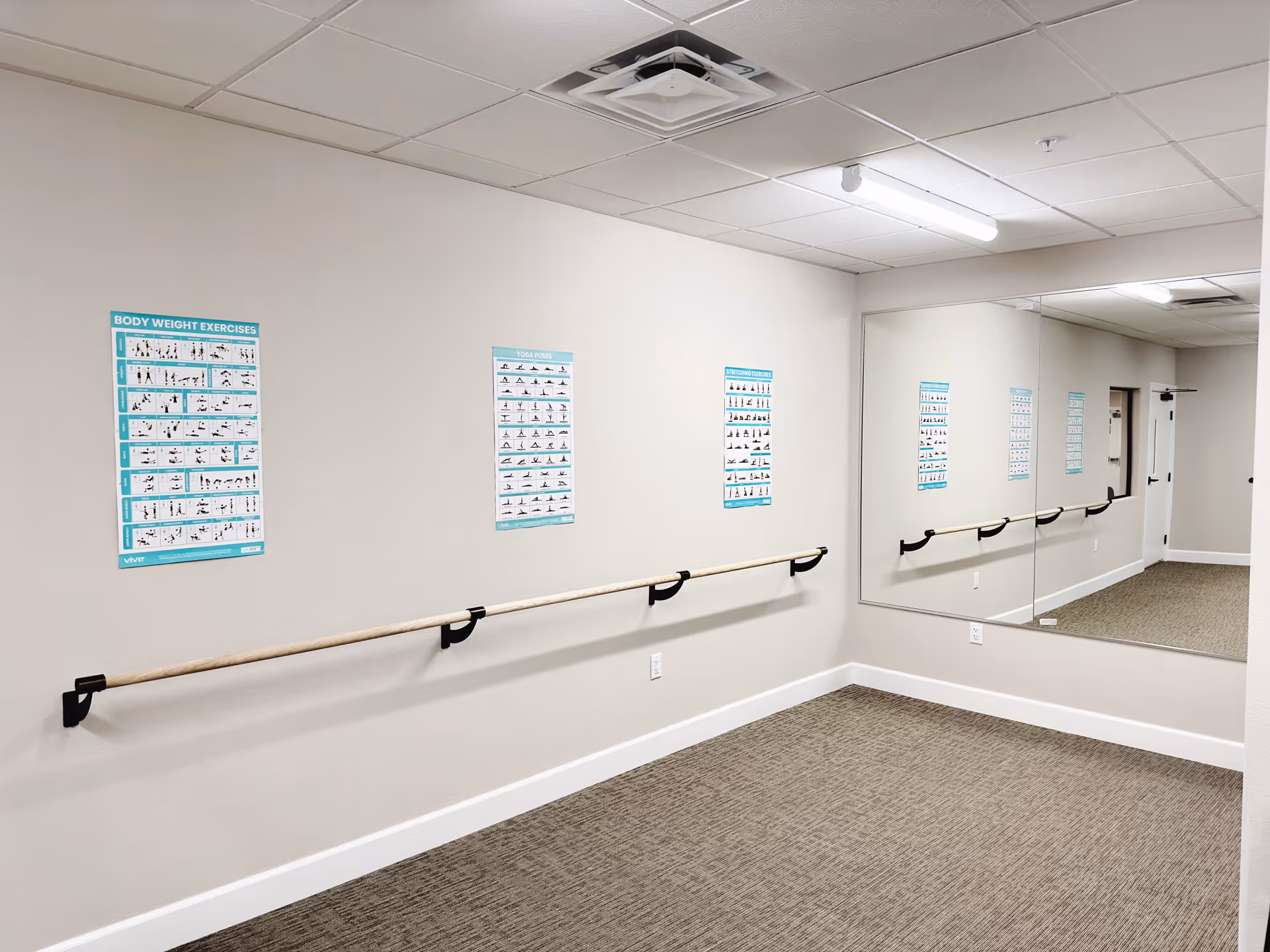 A small exercise room with beige walls and carpeted floor, featuring a long wooden handrail mounted on the wall. Several posters illustrating different exercises such as body weight exercises and yoga poses are displayed above the handrail. A large mirror covers one wall, reflecting the room and posters. The ceiling has fluorescent lighting and a ventilation unit.