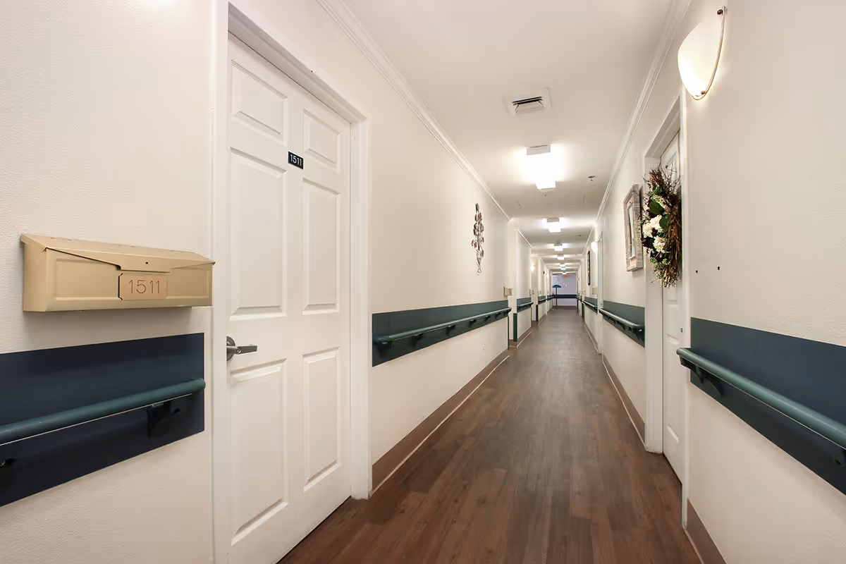 A long, well-lit hallway in an assisted living facility with white walls, dark handrails on both sides, and wooden flooring. Several white doors line the corridor, one marked with the number 1511 and a mailbox beside it. The hallway is decorated with wall art and a wreath on one door.