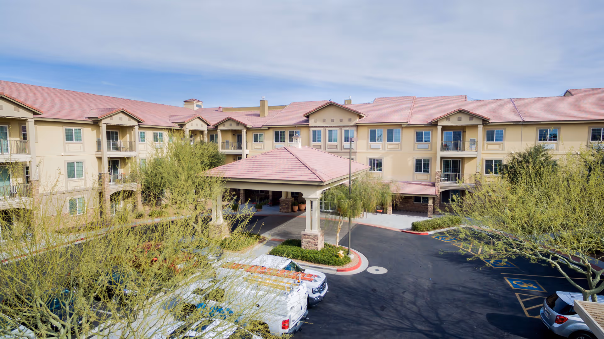 Exterior view of a three-story senior living facility with beige walls and red-tiled roofs. The building has multiple balconies and windows. There is a covered entrance area with columns and a driveway in front. Several trees and parked vehicles are visible in the foreground under a partly cloudy sky.