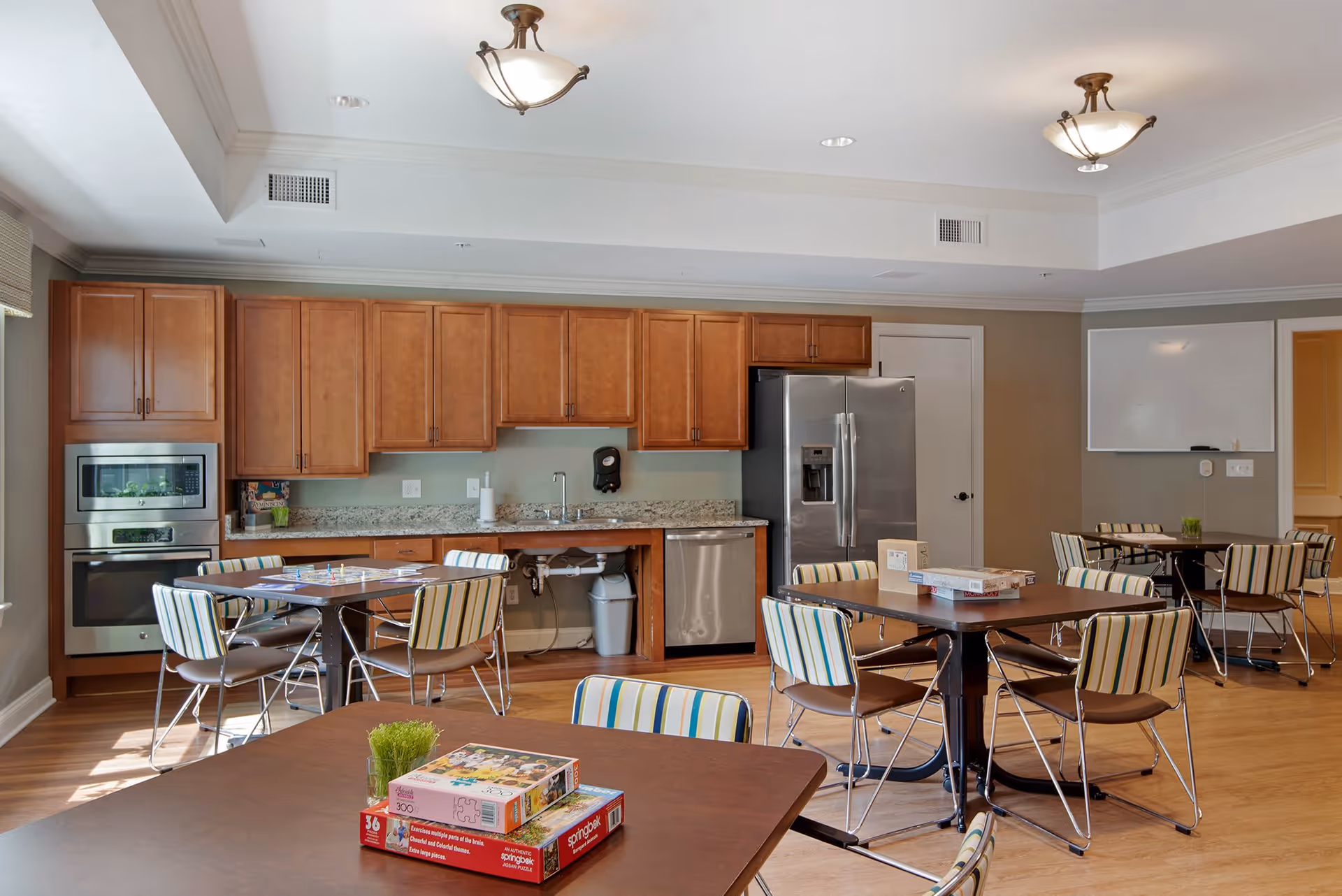 A bright room with a kitchen area featuring wooden cabinets, a stainless steel refrigerator, oven, and dishwasher. Several tables with striped chairs are arranged in the room, with board games and puzzles placed on the tables. The room has wooden flooring and ceiling lights, with a whiteboard on one wall.