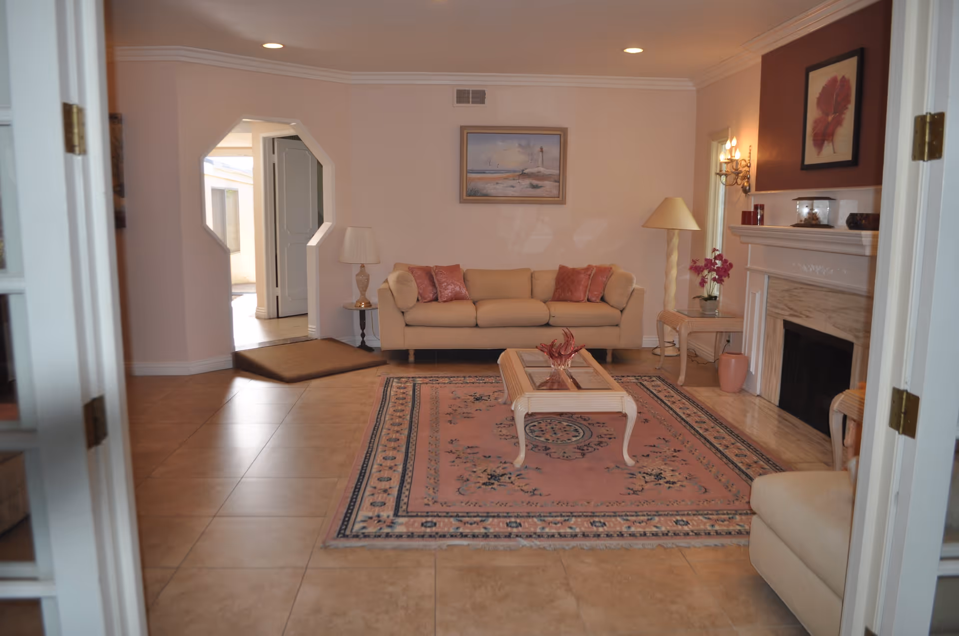 A cozy living room with beige tiled flooring, a light beige sofa with pink cushions, a matching armchair, a glass-top coffee table on a pink patterned rug, a fireplace with a framed leaf artwork above it, and soft lighting from lamps and wall sconces.