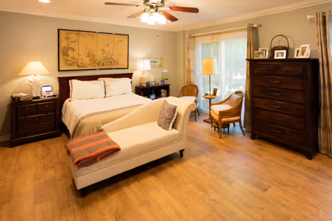 A warmly lit bedroom with a large bed featuring white bedding and pillows, a chaise lounge at the foot of the bed with a folded blanket, two wooden nightstands with lamps on either side of the bed, a wooden dresser with framed photos on top, two chairs and a small round table near a window with curtains, and a ceiling fan with lights overhead.