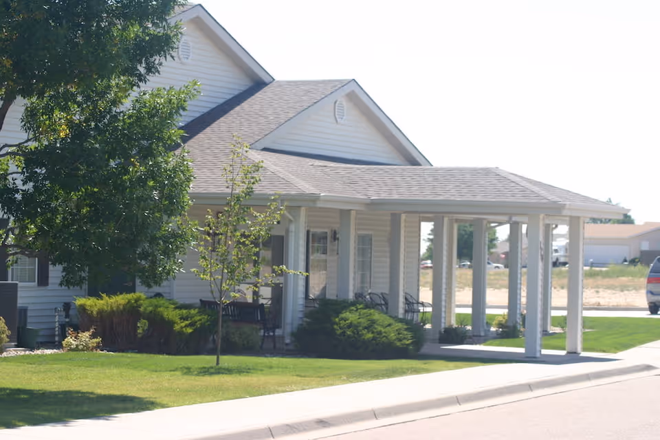 Exterior view of a single-story building with white siding and a covered porch supported by white columns. The porch has several chairs and is surrounded by green bushes and a small tree. A sidewalk and a street are visible in the foreground, with a parked vehicle in the background.