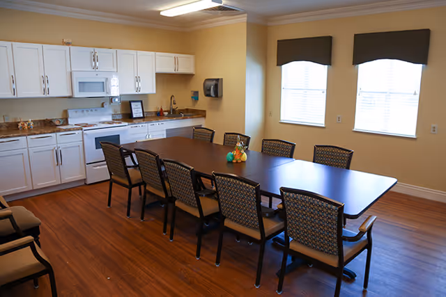 Dining area with a long dark table surrounded by chairs next to a white kitchenette and two windows.