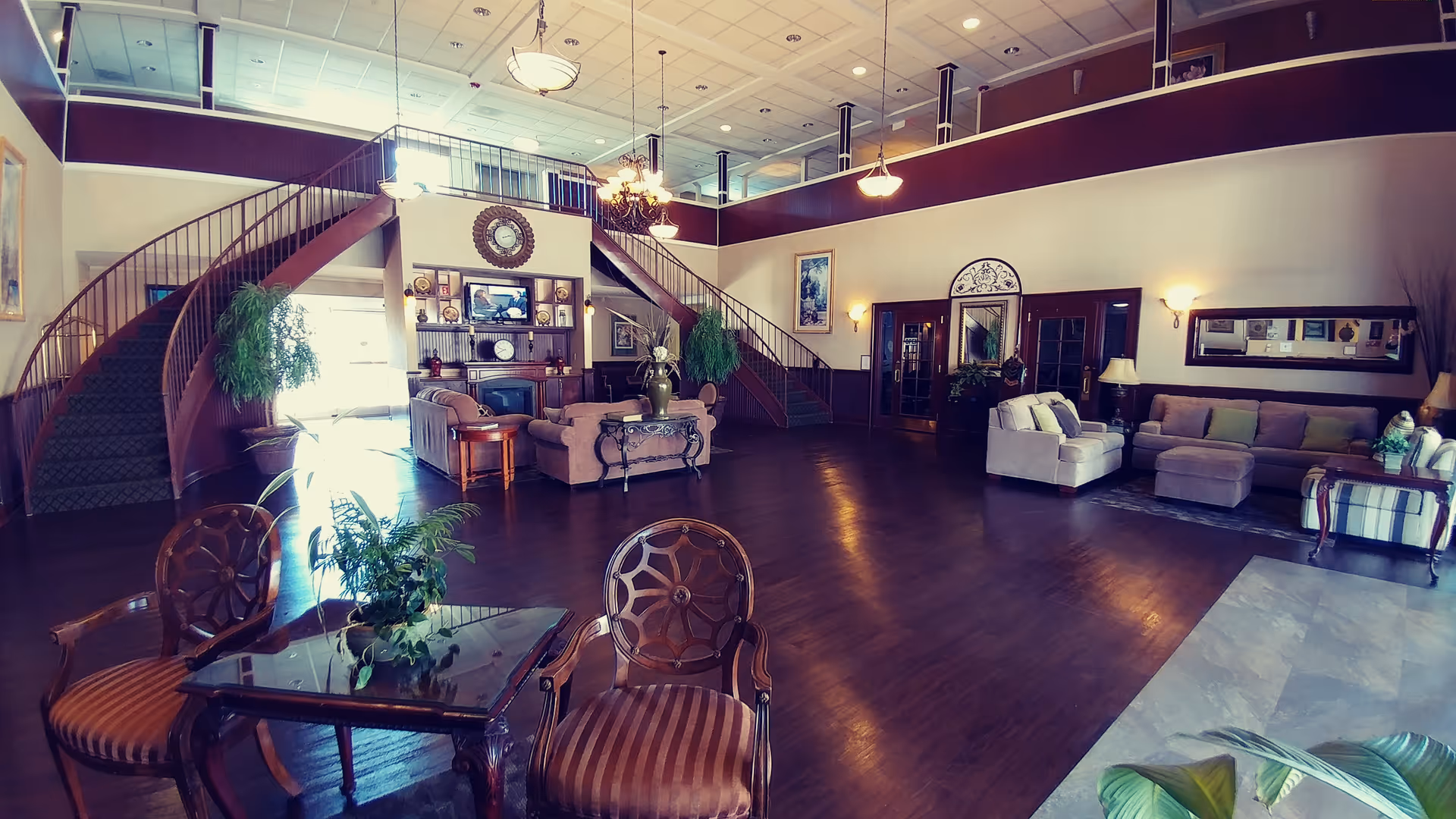 Spacious senior living community common area with high ceilings, two staircases leading to an upper level, multiple seating areas including sofas and armchairs, a fireplace with decorative items, and several plants. The room has wooden flooring and is well-lit with hanging light fixtures and wall sconces.