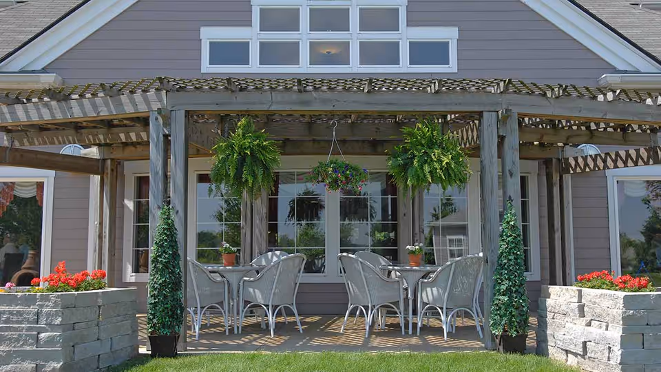 Outdoor patio area with a wooden pergola, two tables each surrounded by four white wicker chairs, hanging green ferns and a flowering plant, stone planters with red flowers on either side, and a building with large windows in the background.