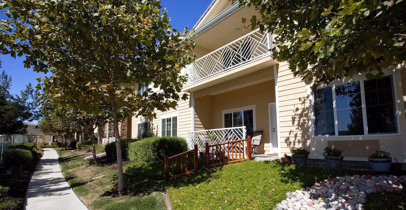 Exterior view of a senior living facility showing a beige two-story building with white railings on balconies and patios. There is a well-maintained lawn with green grass, bushes, and trees lining a concrete walkway under a clear blue sky.