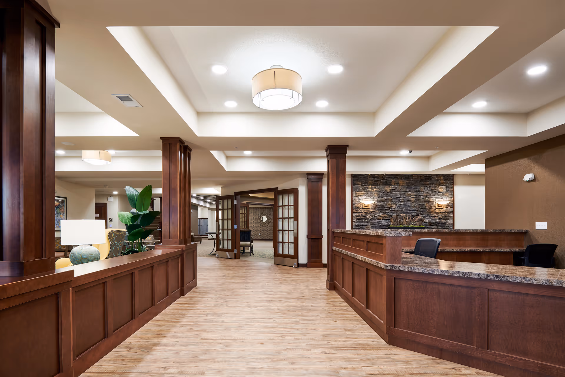 Interior view of a senior living facility reception area with wooden paneling, a stone accent wall with two wall sconces, a granite countertop reception desk with chairs, and a ceiling light fixture. The floor is light wood, and there are glass double doors leading to another room.
