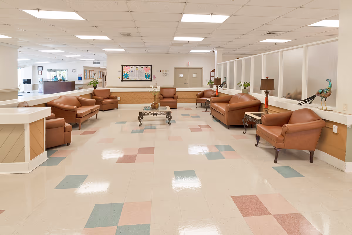 A spacious seating area in a senior living facility with multiple brown leather armchairs and sofas arranged around a glass coffee table. The floor has a patterned tile design in shades of pink, green, and beige. There are decorative items including a peacock sculpture and table lamps on side tables. The background shows a reception desk and a bulletin board on the wall.