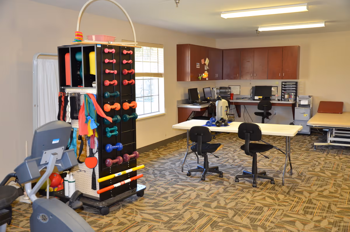A rehabilitation or physical therapy room with exercise equipment including dumbbells, resistance bands, and stationary exercise machines. There are two black chairs at a white table, a padded examination or therapy table, and a desk area with computers and office supplies. The room has patterned carpet flooring and a window letting in natural light.