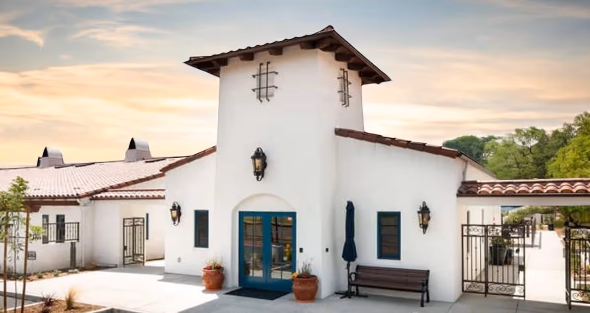 Exterior view of a white building with a red tile roof and a central tower-like structure. The building has blue doors and windows, black wrought iron gates, outdoor wall lanterns, a bench, and potted plants near the entrance. The sky is partly cloudy with a warm sunset glow.