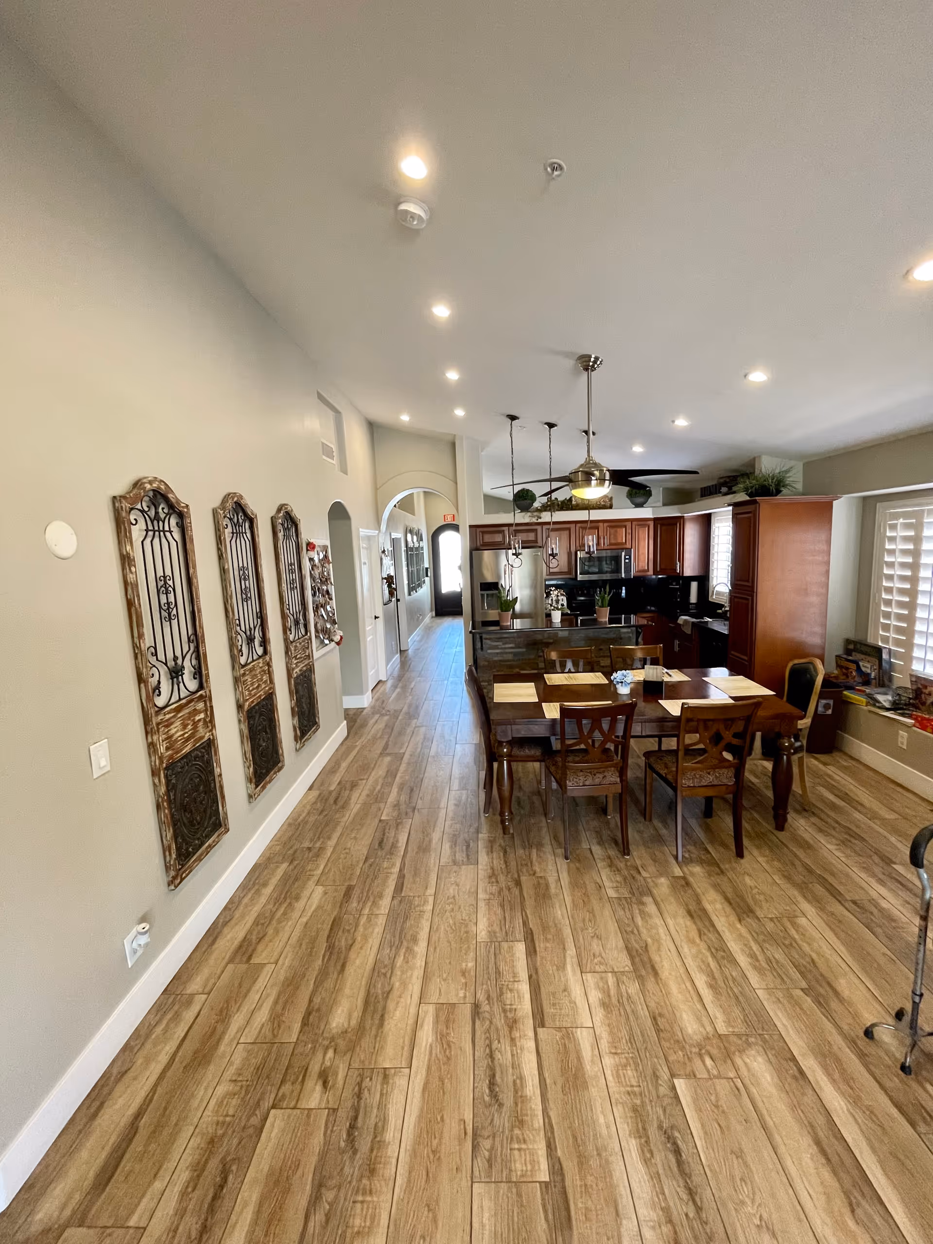 Interior view of a senior living facility showing a long hallway with wood-look flooring leading to a kitchen and dining area. The kitchen has wooden cabinets, a black countertop, and stainless steel appliances. A dining table with chairs is set with placemats. Decorative wall panels are mounted on the left wall, and there is a window with plantation shutters on the right.