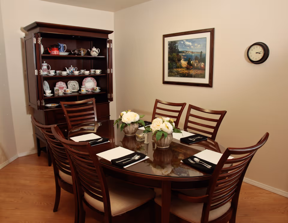 A dining room with a wooden table set for six people, each place setting includes a white placemat, black napkin, fork, and spoon. Two small flower arrangements are placed in the center of the table. Behind the table is a wooden cabinet displaying various teapots, cups, and plates. A framed landscape painting hangs on the wall, and a round clock shows the time as 1:10.