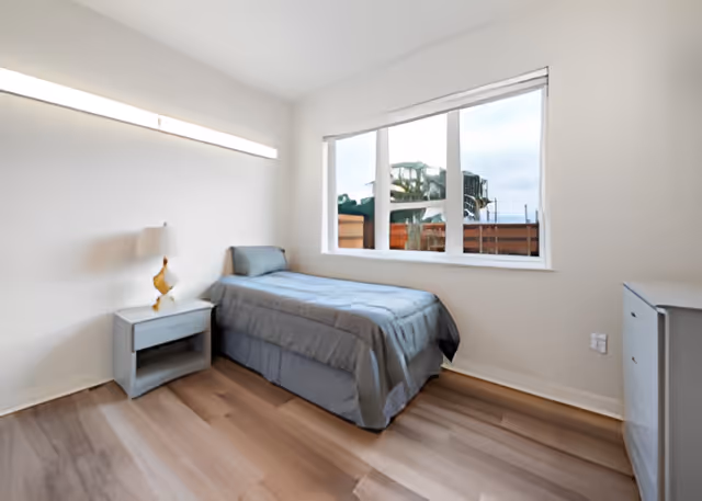 A simple, clean bedroom in a senior living facility with a single bed covered in a gray bedspread, a small nightstand with a lamp, a large window showing an outdoor view, and light-colored wooden flooring.