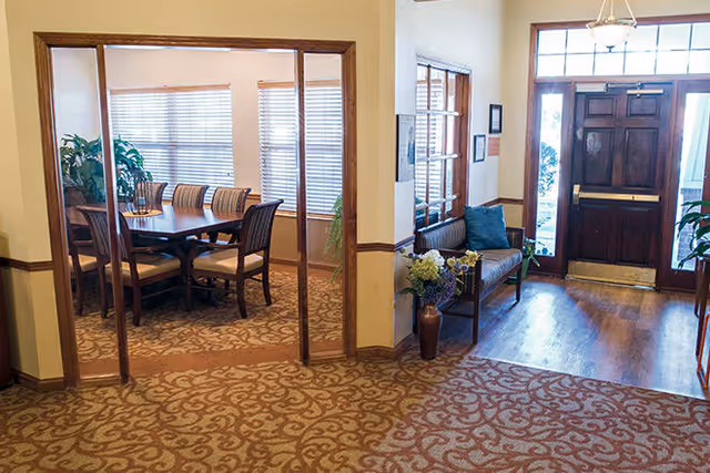 A facility entrance foyer with a wooden front door and bench opening into a dining room with a table and chairs.