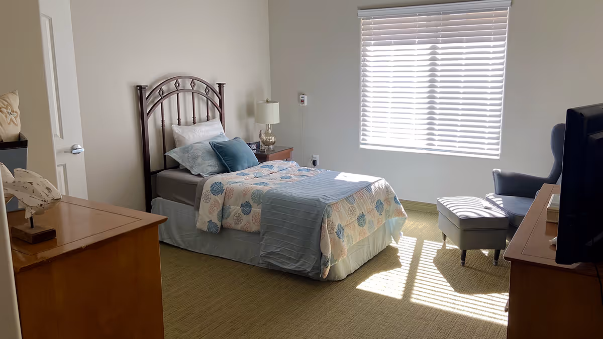A well-lit bedroom with a single bed featuring a metal headboard, a bedside table with a lamp, a cushioned armchair with an ottoman near a window with blinds, a wooden dresser, and a television on a stand.