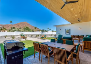 Covered outdoor patio with multiple dining tables and chairs, lounge seating, a barbecue grill, artificial turf, and a mountain backdrop.