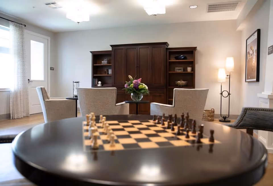 Cozy common room with a chessboard on a round table in the foreground, upholstered chairs facing a wooden cabinet and bookshelf, and a standing lamp.