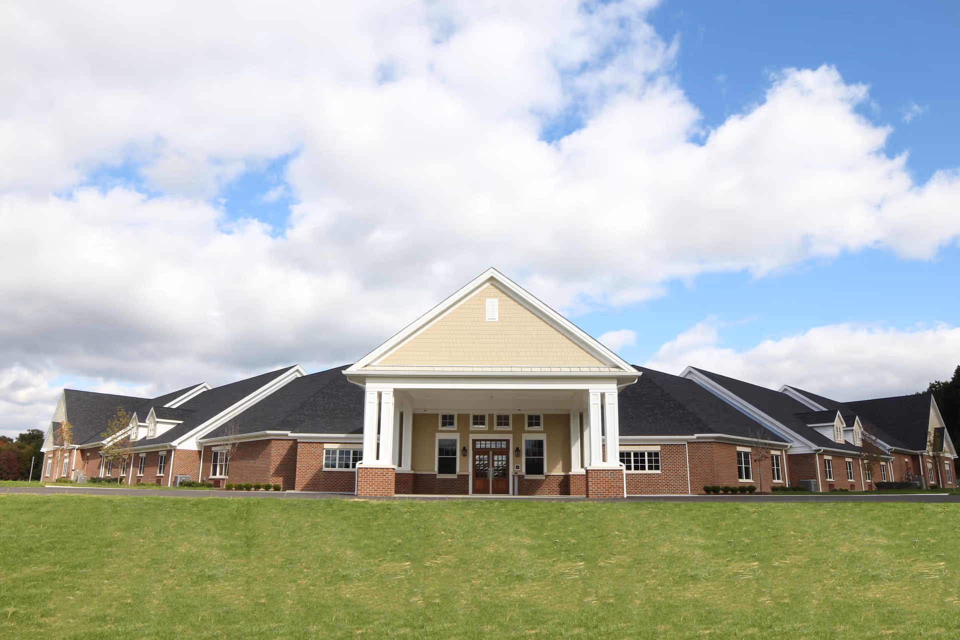 Front exterior of a single-story brick assisted living building with a columned entrance and lawn under a partly cloudy sky.