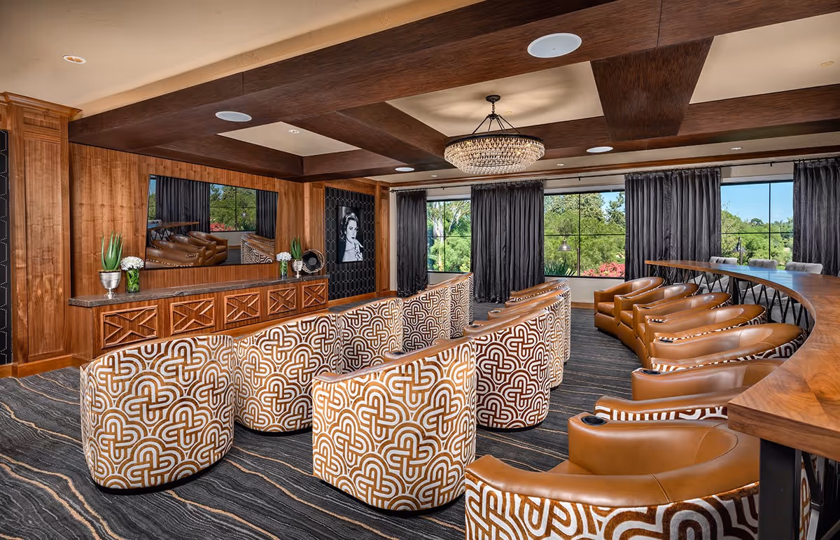 Upscale communal lounge with patterned upholstered swivel chairs, leather armchairs along a curved wooden bar, large windows and a central chandelier.