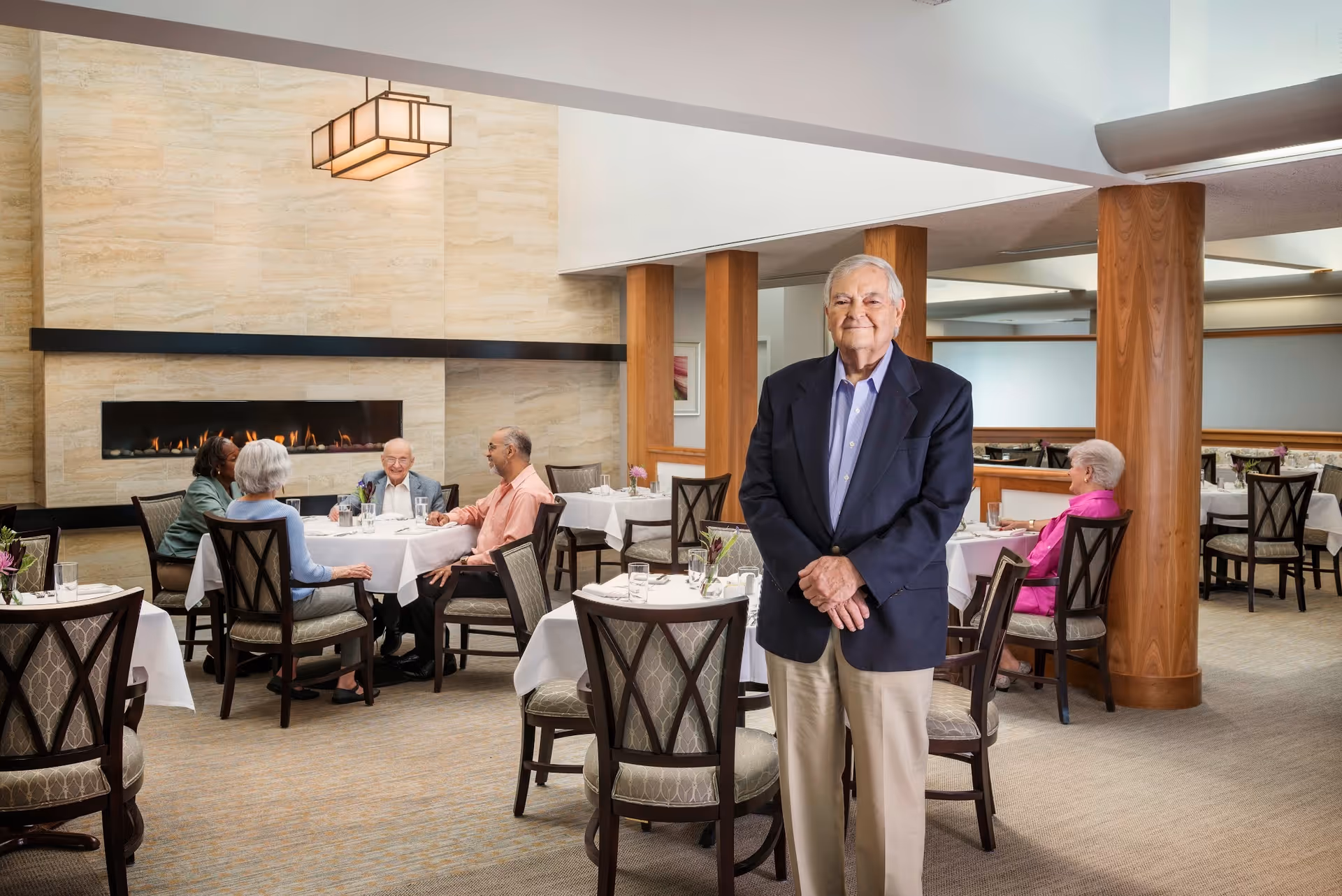 A dining room in a retirement community with several elderly people seated at tables covered with white tablecloths. A man in a dark blazer and beige pants stands in the foreground smiling. The room features a modern fireplace on the left wall and wooden columns throughout.