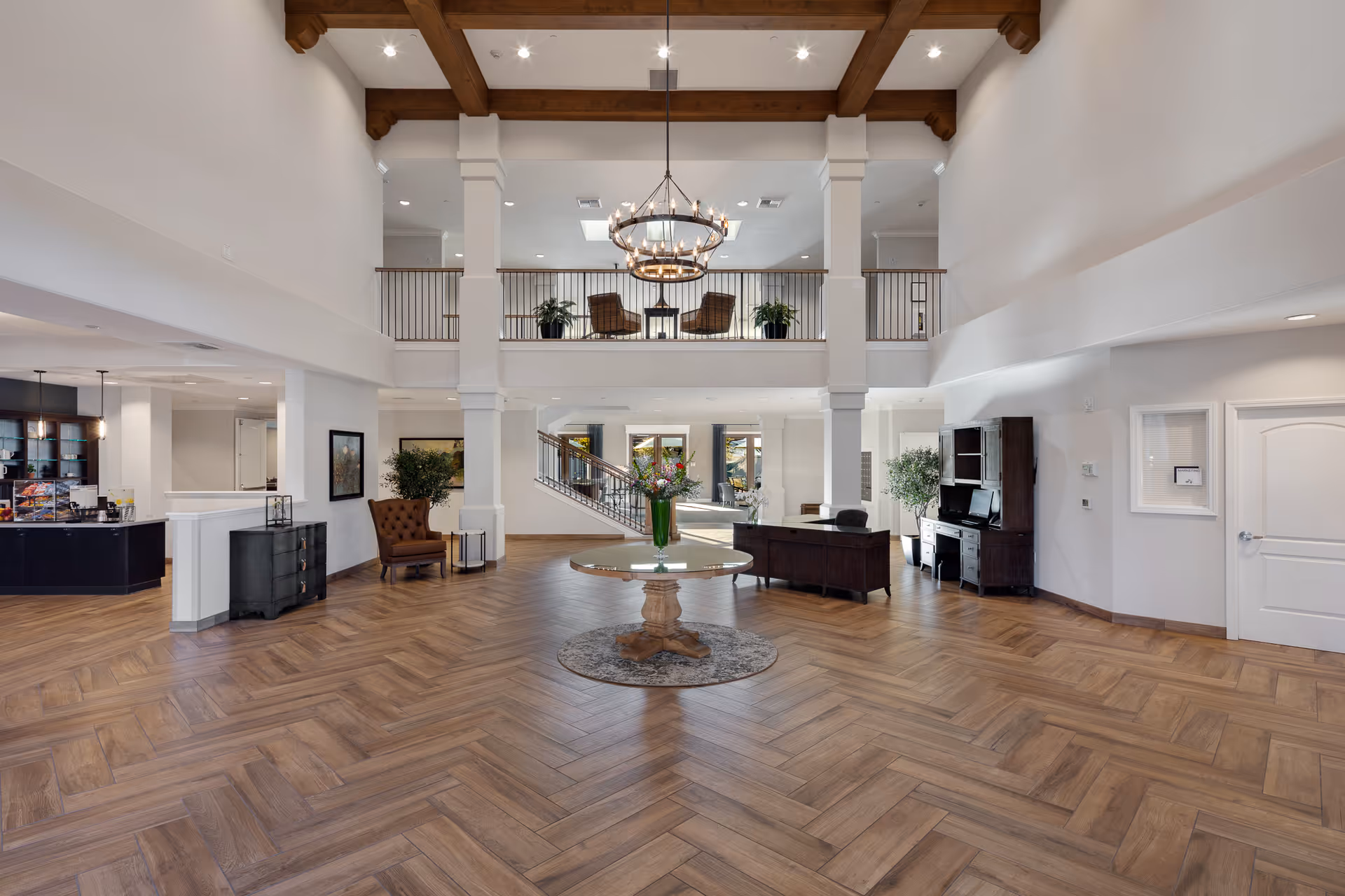 Spacious and elegant senior living facility lobby with high ceilings, wooden beams, a large chandelier, and a round table with a flower arrangement in the center. The room features wood-patterned flooring, comfortable seating areas, plants, and a reception desk. A staircase and balcony with chairs and plants are visible in the background.