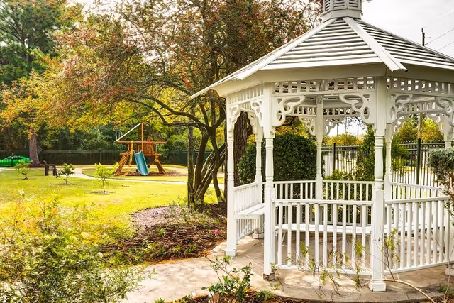 White wooden gazebo in a landscaped outdoor area with trees and a children's playset visible in the background.