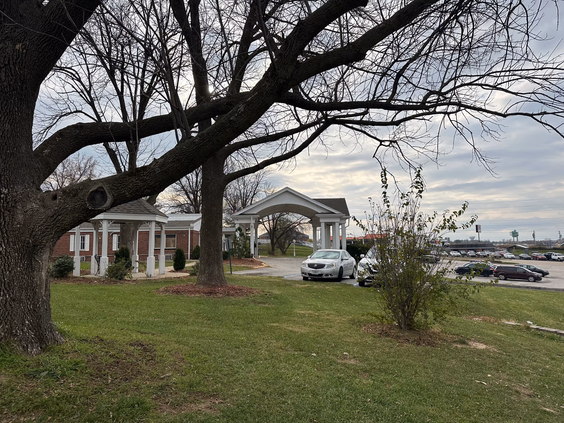 Exterior view of Aspen Point Health and Rehabilitation showing a brick building with white columns and a covered entrance. There are large leafless trees and a grassy lawn in the foreground, with a parking lot and several cars in the background under a cloudy sky.