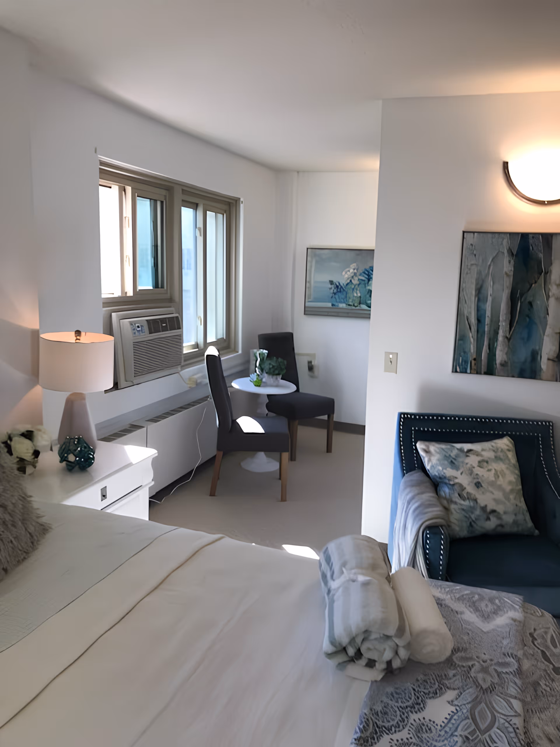 Bright furnished bedroom in a senior living unit with a bed, armchair, small table with two chairs, and a window air conditioner.