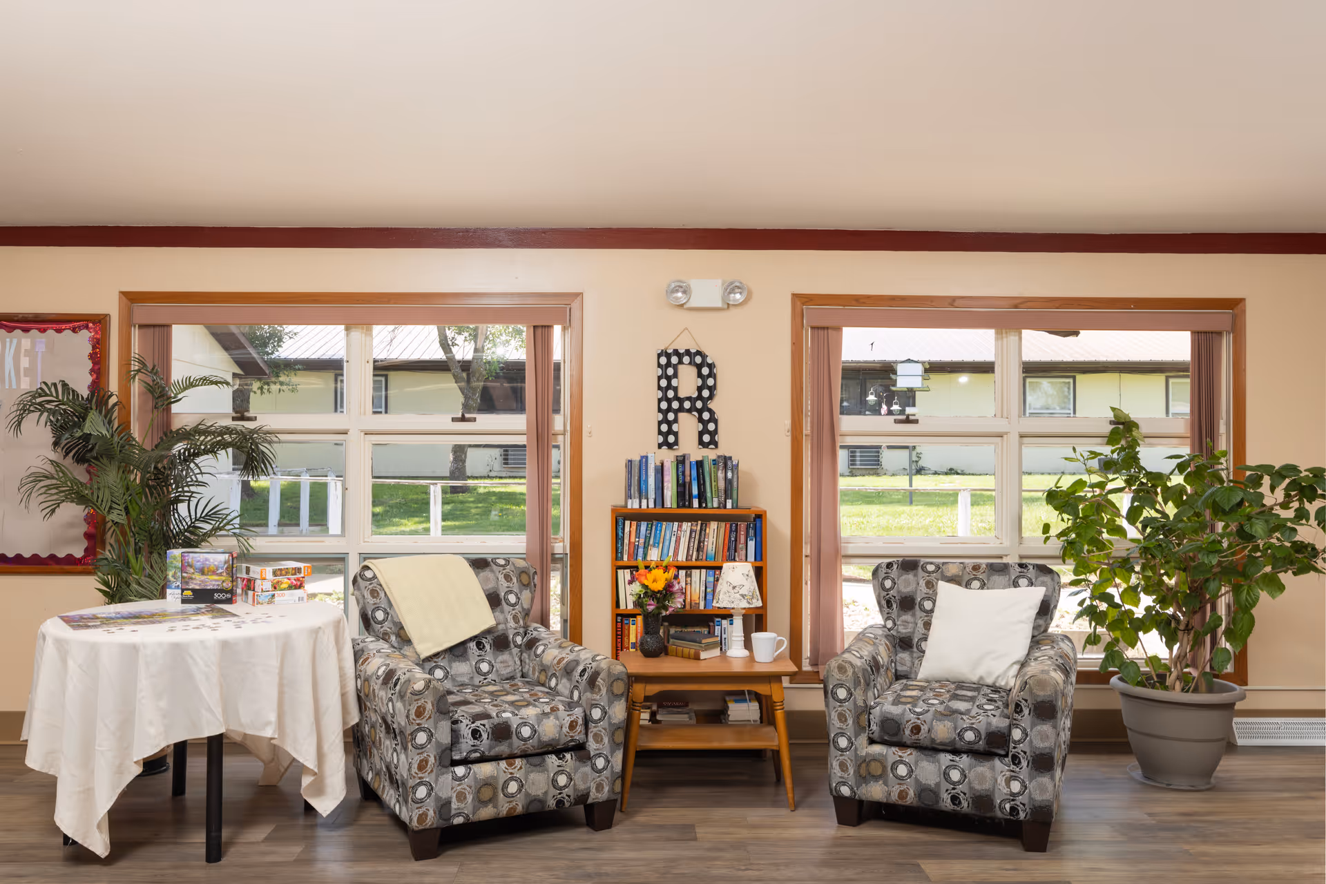 Two patterned armchairs flanking a small bookshelf and table in a bright communal sitting area with plants and large windows.