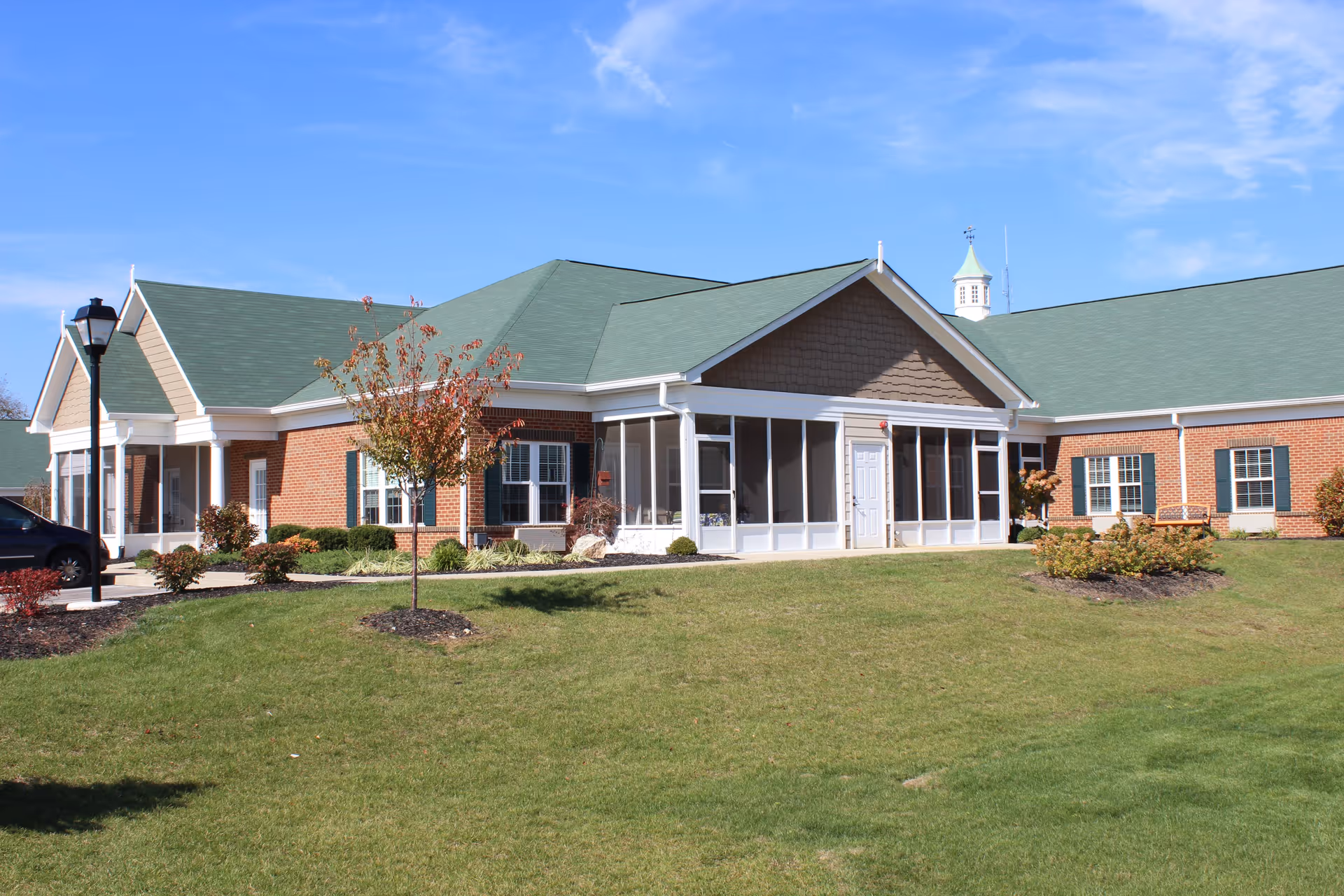 Exterior view of a single-story senior living facility building with a green roof, brick walls, and white trim. The building has multiple windows with green shutters, a screened porch area, and a small tree and landscaped bushes in front. The sky is clear and blue.