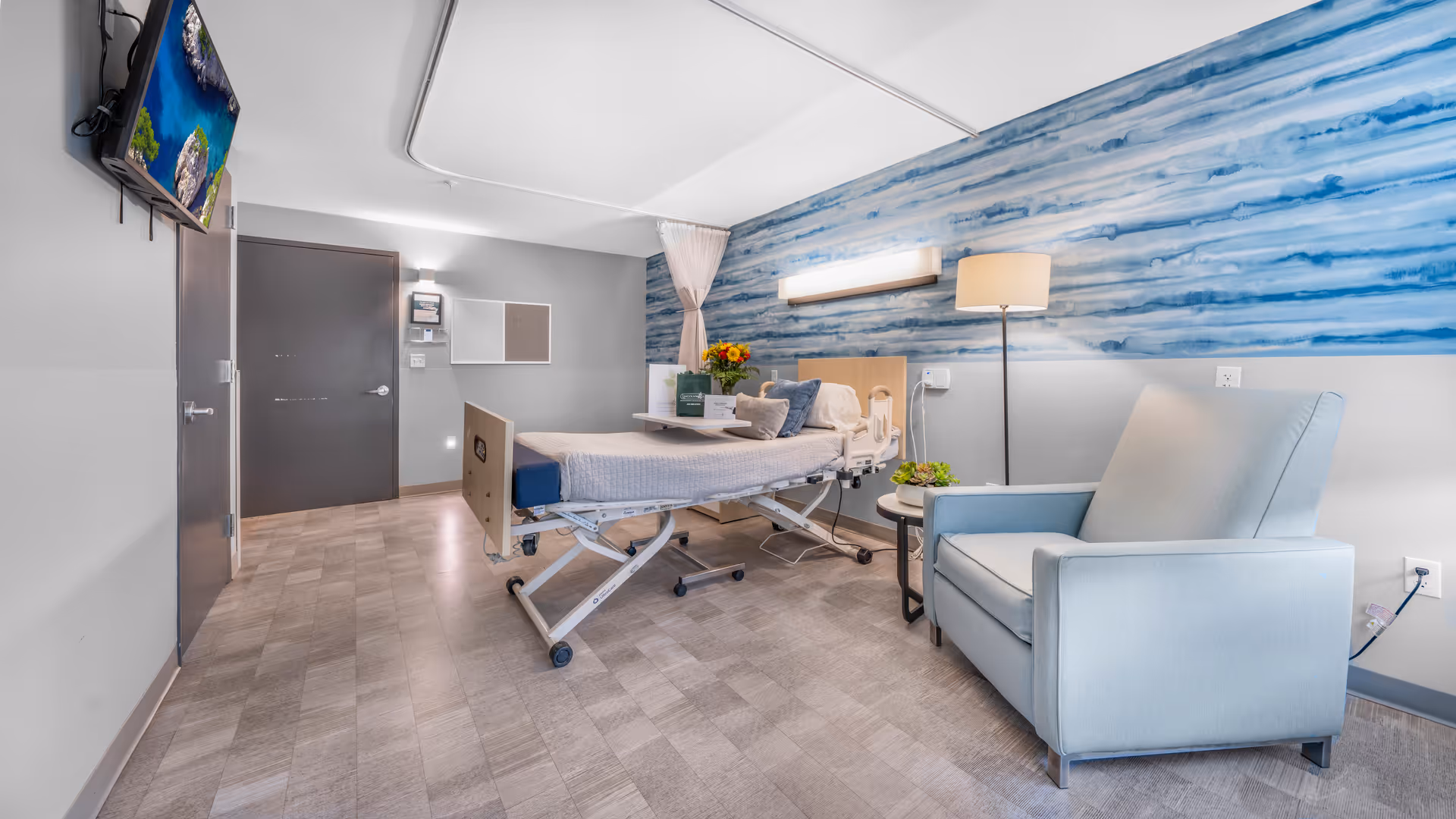 A bright rehabilitation patient room with a hospital bed, blue accent wall, armchair, wall-mounted TV and bedside table.