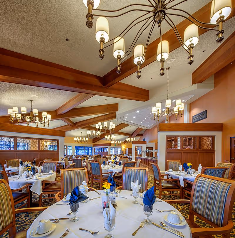 Spacious dining room with multiple round tables covered in white tablecloths, each set with plates, silverware, glasses, and napkins folded in decorative shapes. The room features wooden beams on the ceiling, multiple chandeliers with warm lighting, and large windows along the walls. Chairs have striped upholstery and the carpet has an intricate pattern.