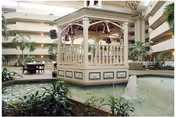 Indoor atrium area with a central gazebo surrounded by a water feature with a small fountain, plants, and seating areas. The space is enclosed by multiple floors with balconies overlooking the atrium.