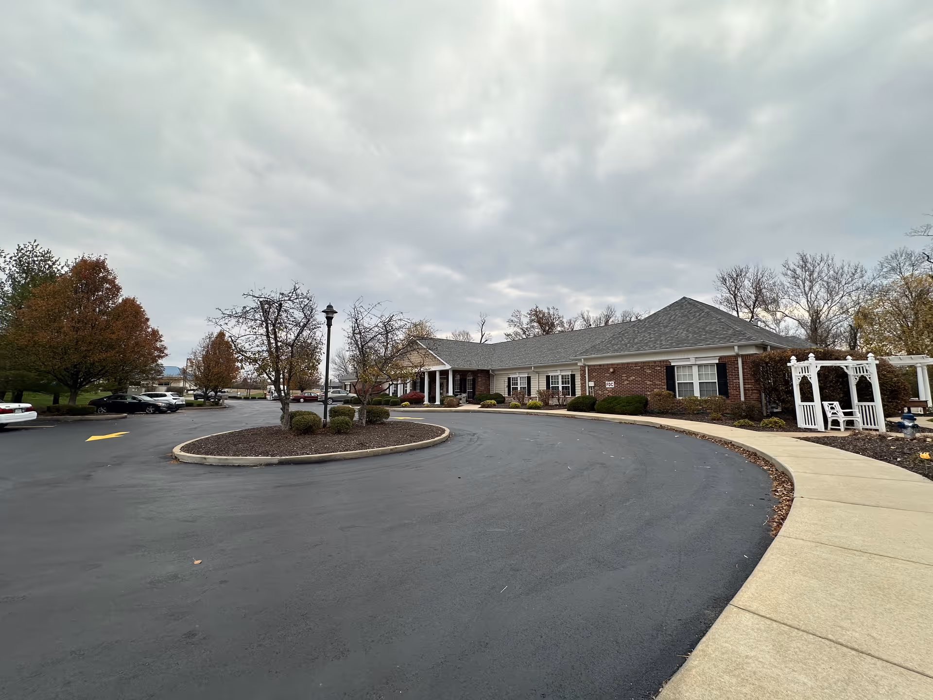 Exterior view of a single-story brick building with a gray roof, surrounded by a curved driveway and landscaped areas with bushes and trees under a cloudy sky. There is a white arbor with chairs on the right side near the sidewalk.