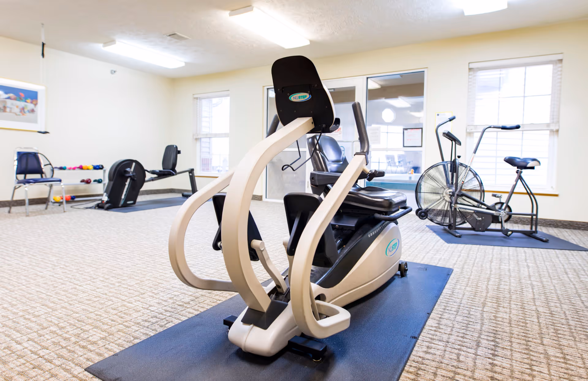 A senior living facility exercise room with various fitness equipment including a NuStep recumbent cross trainer in the foreground, a stationary exercise bike, a rowing machine, and a rack of colorful dumbbells. The room has beige carpet, cream-colored walls, two windows with blinds, and fluorescent ceiling lights.