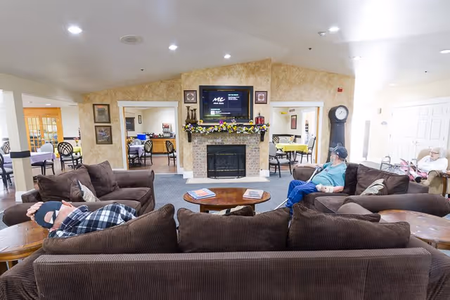 A senior living facility common area with brown sofas arranged around a wooden coffee table. Two elderly men are seated on the sofas, one resting and the other looking towards the fireplace. The fireplace has a TV mounted above it and is decorated with flowers. In the background, there are dining tables and chairs with yellow and purple tablecloths, and another elderly person is seated near the right side of the room.