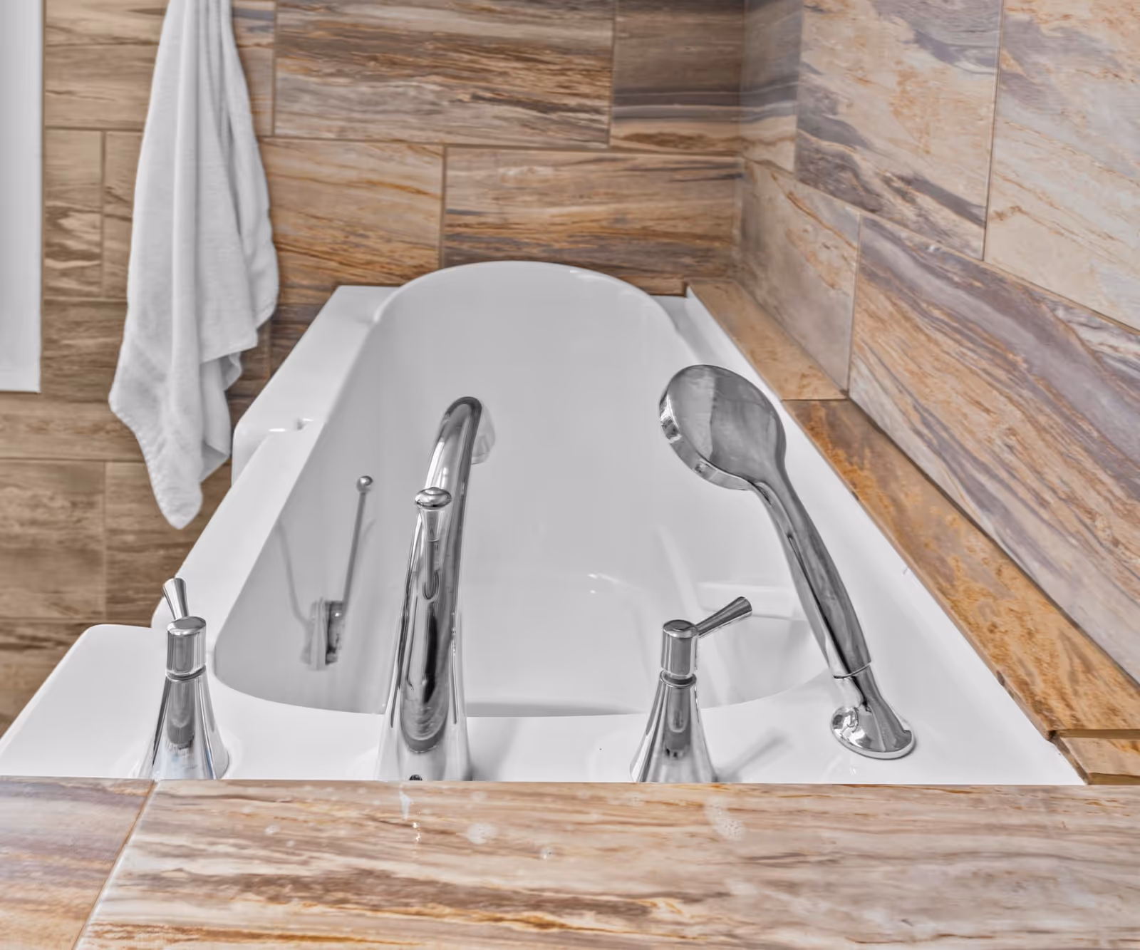 Close-up view of a white bathtub with chrome faucet and handheld showerhead, surrounded by beige and brown marble tiles. A white towel hangs on a hook on the wall to the left.