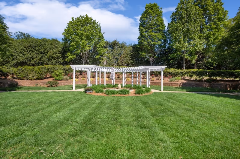 A wide green lawn with a white pergola at center surrounded by trees and shrubs under a blue sky.